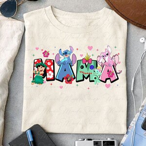 May include: A beige t-shirt with a colorful graphic design featuring the word "MAMA" spelled out with cartoon characters from the Disney movie Lilo & Stitch. The characters include Stitch, Angel, and Scrump. The letters are decorated with flowers and hearts.