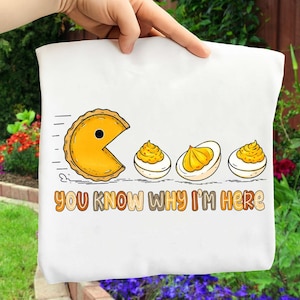 May include: White t-shirt featuring a Pac-Man-inspired pie character chasing deviled eggs. The text "YOU KNOW WHY I'M HERE" is printed in brown and orange. The design is a playful take on a classic video game.