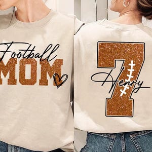 May include: Two beige sweatshirts with a football theme. One sweatshirt has the text "Football Mom" in brown glitter letters with a heart. The other sweatshirt has a brown glitter number "7" with the name "Henry" written in cursive below it.