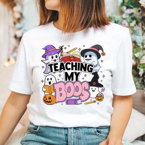 May include: White t-shirt with a graphic of ghosts and the text "Teaching My Boo's". The ghosts are wearing witch hats and holding books. There are also stars and pencils in the design.