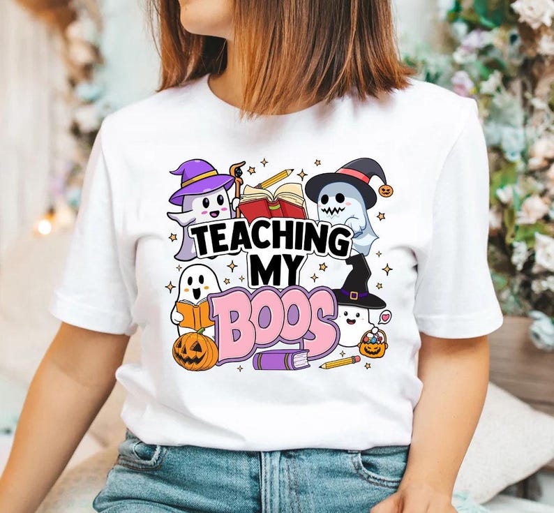 May include: White t-shirt with a graphic of ghosts and the text "Teaching My Boo's". The ghosts are wearing witch hats and holding books. There are also stars and pencils in the design.