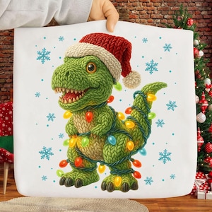 May include: A white tote bag featuring a green knitted dinosaur wearing a red Santa hat and wrapped in colorful Christmas lights. Blue snowflakes are scattered around the dinosaur, creating a festive holiday theme.