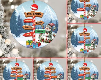 May include: A Christmas ornament featuring a snowman with a red hat and a signpost with the names "The Smith", "Sophia", "David", "Amelia", "Lucas", "Henry", and "Max". The ornament is decorated with a winter scene and a red and white candy cane.