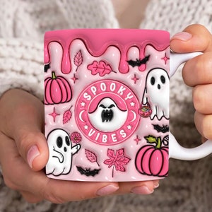 May include: Pink ceramic mug with a white handle. The mug has a dripping pink glaze and features a spooky Halloween theme with ghosts, pumpkins, bats, and leaves. The text "Spooky Vibes" is in a circle with a ghost inside.