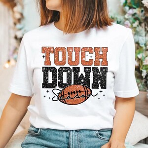 May include: White t-shirt with the text "Touchdown Season" in glittery orange and black letters. A brown and orange football graphic is below the text.
