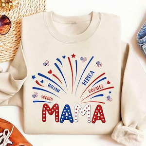May include: A cream-coloured sweatshirt featuring a celebratory design. The word "MAMA" is in red and white polka-dot letters. Above, a firework design in red, white, and blue includes names such as William, Sophia, Rebeca, and Gonzalez.
