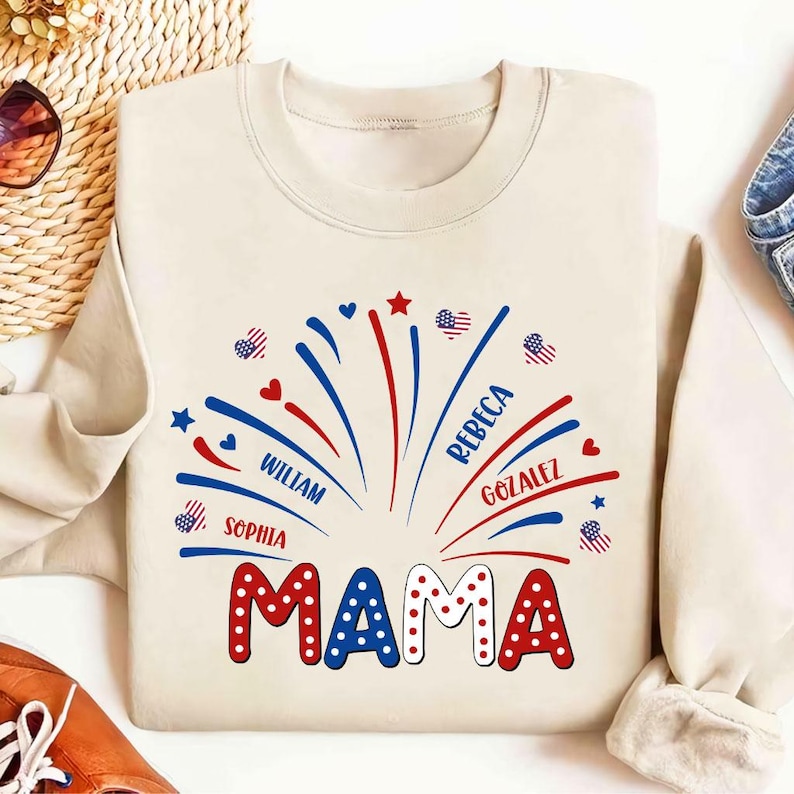 May include: A cream-coloured sweatshirt featuring a celebratory design. The word "MAMA" is in red and white polka-dot letters. Above, a firework design in red, white, and blue includes names such as William, Sophia, Rebeca, and Gonzalez.