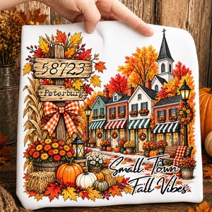 Può includere: Una felpa bianca con una scena autunnale colorata, un cartello in legno con scritto 58723 Peterburg, un fiocco a quadri, zucche, balle di fieno ed edifici con un campanile. Il testo Small Town Fall Vibes è stampato in corsivo nero.