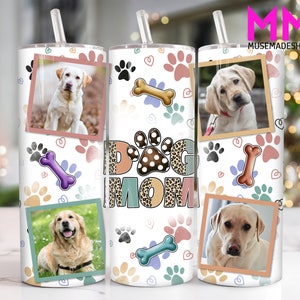 Puede incluir: Un vaso blanco con un diseño de "Dog Mom". El vaso tiene un fondo blanco con huellas de patas, corazones y huesos. El texto "Dog Mom" está en marrón, negro y blanco con un diseño de estampado de leopardo. Hay tres fotos de perros en el vaso.