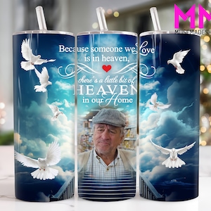 May include: A white tumbler with a blue and white cloudy sky background. The tumbler has a quote that reads "Because someone we love is in heaven, there's a little bit of heaven in our home". There is a photo of an older man in the center of the tumbler.