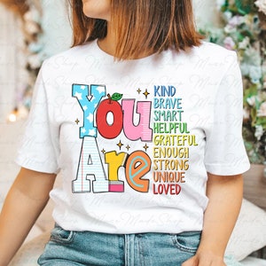以下が含まれることがあります： 赤いリンゴと鉛筆が描かれた「You Are」というカラフルなグラフィックがプリントされた白いTシャツ。グラフィックには、「Kind、Brave、Smart、Helpful、Grateful、Enough、Strong、Unique、Loved」という単語も含まれています。