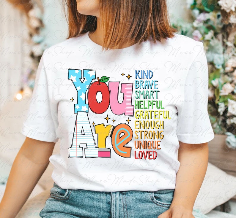 以下が含まれることがあります： 赤いリンゴと鉛筆が描かれた「You Are」というカラフルなグラフィックがプリントされた白いTシャツ。グラフィックには、「Kind、Brave、Smart、Helpful、Grateful、Enough、Strong、Unique、Loved」という単語も含まれています。