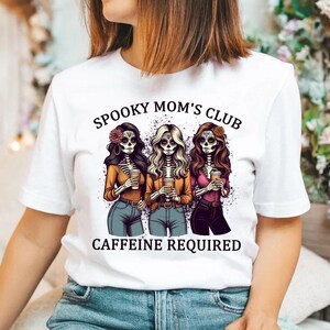 May include: White t-shirt with a graphic of three skeleton women holding coffee cups. The text "Spooky Mom's Club" and "Caffeine Required" is printed on the shirt.