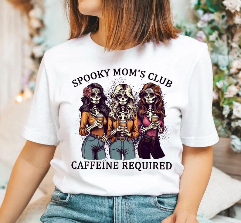 May include: White t-shirt with a graphic of three skeleton women holding coffee cups. The text "Spooky Mom's Club" and "Caffeine Required" is printed on the shirt.