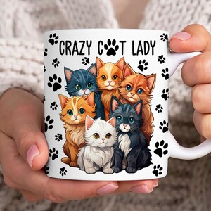 May include: White ceramic mug with a black paw print pattern and the text "Crazy Cat Lady". The mug features a cartoon illustration of six cats in various colors and poses.