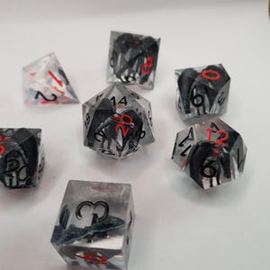 May include: Set of seven polyhedral dice. The dice are clear with black and gray internal designs and red numbering. The dice are designed for tabletop role-playing games.