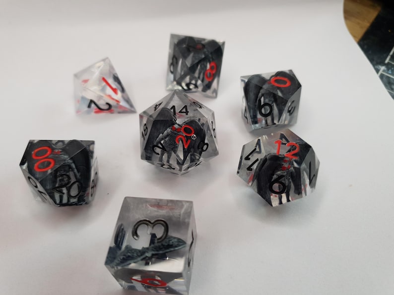 May include: Set of seven polyhedral dice. The dice are clear with black and gray internal designs and red numbering. The dice are designed for tabletop role-playing games.