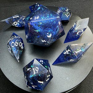 Puede incluir: Un conjunto de siete dados poliédricos de varias formas, con un diseño cósmico azul profundo y brillante. Los dados tienen numeración blanca y están diseñados para juegos de rol de mesa.
