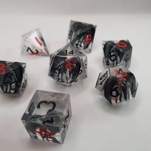 May include: A collection of clear, multi-sided dice with black and red accents. The dice feature black designs within the clear resin, with red numbers. The dice are scattered on a white surface, suitable for tabletop gaming.