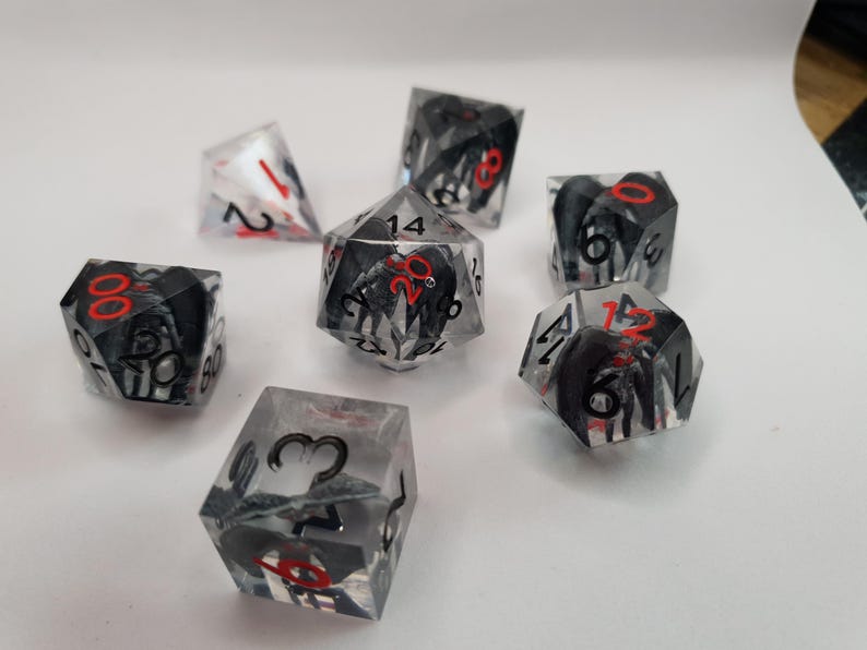 May include: A collection of clear, multi-sided dice with black and red accents. The dice feature black designs within the clear resin, with red numbers. The dice are scattered on a white surface, suitable for tabletop gaming.
