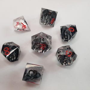 May include: A collection of seven polyhedral dice. The dice are clear with black and gray internal designs and red numbering. The dice are arranged on a white surface.