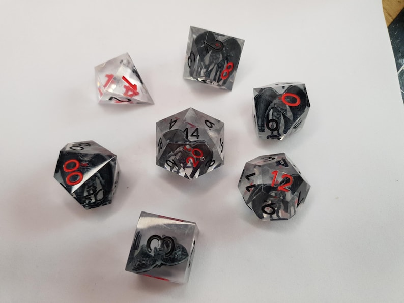May include: A collection of seven polyhedral dice. The dice are clear with black and gray internal designs and red numbering. The dice are arranged on a white surface.