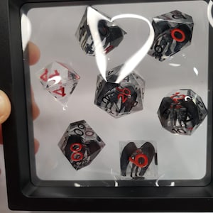 May include: A set of seven polyhedral dice displayed in a black shadow box. The dice are clear with black and red accents, featuring a creature design. One die is clear with red numbers.