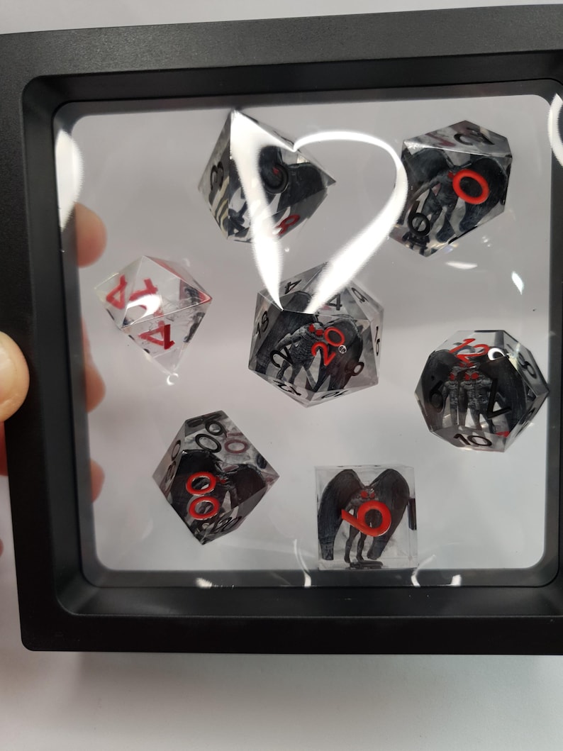 May include: A set of seven polyhedral dice displayed in a black shadow box. The dice are clear with black and red accents, featuring a creature design. One die is clear with red numbers.