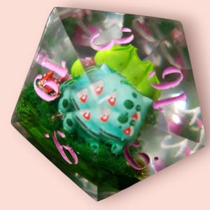 May include: A clear resin die with a teal Bulbasaur figure and red details, set in green moss. The die has pink numbers and decorative elements. A unique collectible item, the die is a fun accessory.