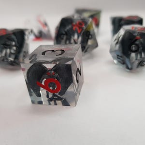 May include: Set of polyhedral dice with a black and red design encased in clear resin. The dice feature a detailed illustration of a creature with wings and red eyes. Ideal for tabletop games.