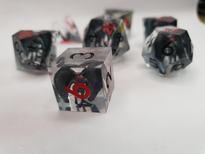 May include: Set of polyhedral dice with a black and red design encased in clear resin. The dice feature a detailed illustration of a creature with wings and red eyes. Ideal for tabletop games.