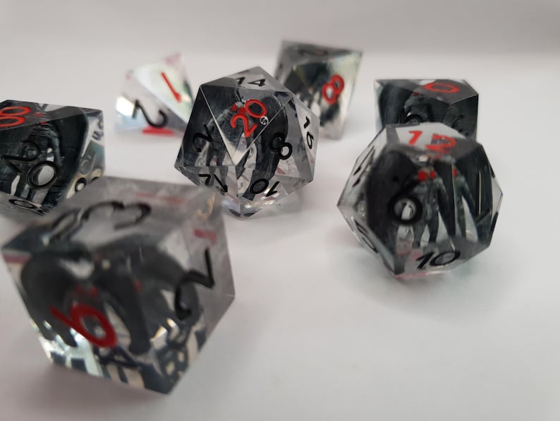 May include: A collection of polyhedral dice with clear resin bodies. Each die features a black and white design inside, with red numbers. The dice are scattered on a white surface, ideal for tabletop gaming.