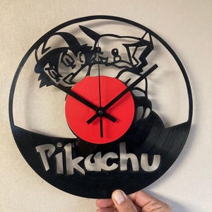 May include: A black vinyl record clock featuring a cut-out design of a character and the word "Pikachu". The clock has a red center and black hands. The clock is held by a hand.