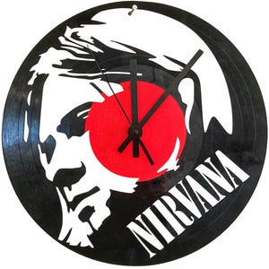 Peut inclure: Une horloge en vinyle noir avec une silhouette blanche d'un visage et le mot "NIRVANA" en blanc. L'horloge a un centre rouge et des aiguilles noires.