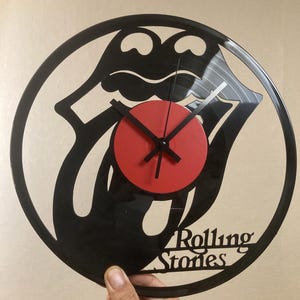 Könnte beinhalten: Schwarze Vinyl-Schallplattenuhr mit dem ikonischen Zungen- und Lippen-Logo der Rolling Stones. Die Uhr hat eine rote Mitte und schwarze Zeiger, mit dem Namen der Band am unteren Rand ausgeschnitten. Ein einzigartiges Dekorationsobjekt.