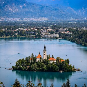 Può includere: Una chiesa bianca con tetto rosso si trova su una piccola isola in un lago. L'isola è ricoperta di alberi verdi e il lago è circondato da montagne.