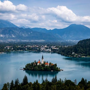 Könnte beinhalten: Eine weiße Kirche mit einem hohen Kirchturm steht auf einer kleinen Insel in einem See. Der See ist umgeben von üppigem Grün und Bergen in der Ferne. Der Himmel ist blau mit weißen Wolken.
