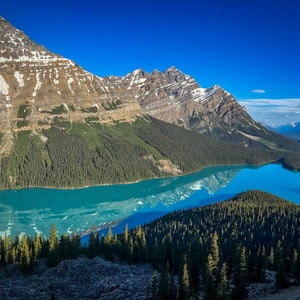 Peut inclure: Vue panoramique d'un lac turquoise niché entre des montagnes enneigées et une forêt dense. L'eau reflète le paysage environnant, créant un effet miroir.