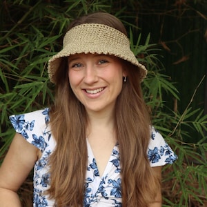 May include: A woman wearing a beige crochet sun visor with a wide brim. She is smiling and has long brown hair. She is wearing a white and blue floral dress.