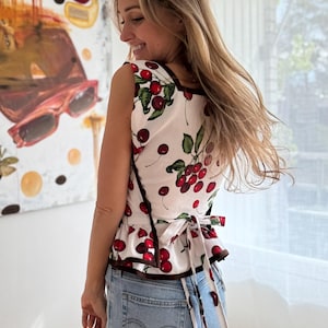 May include: A sleeveless white top with a cherry print, featuring red cherries and green leaves. The top has a brown trim and a tie-back detail. The person is wearing light blue denim jeans.