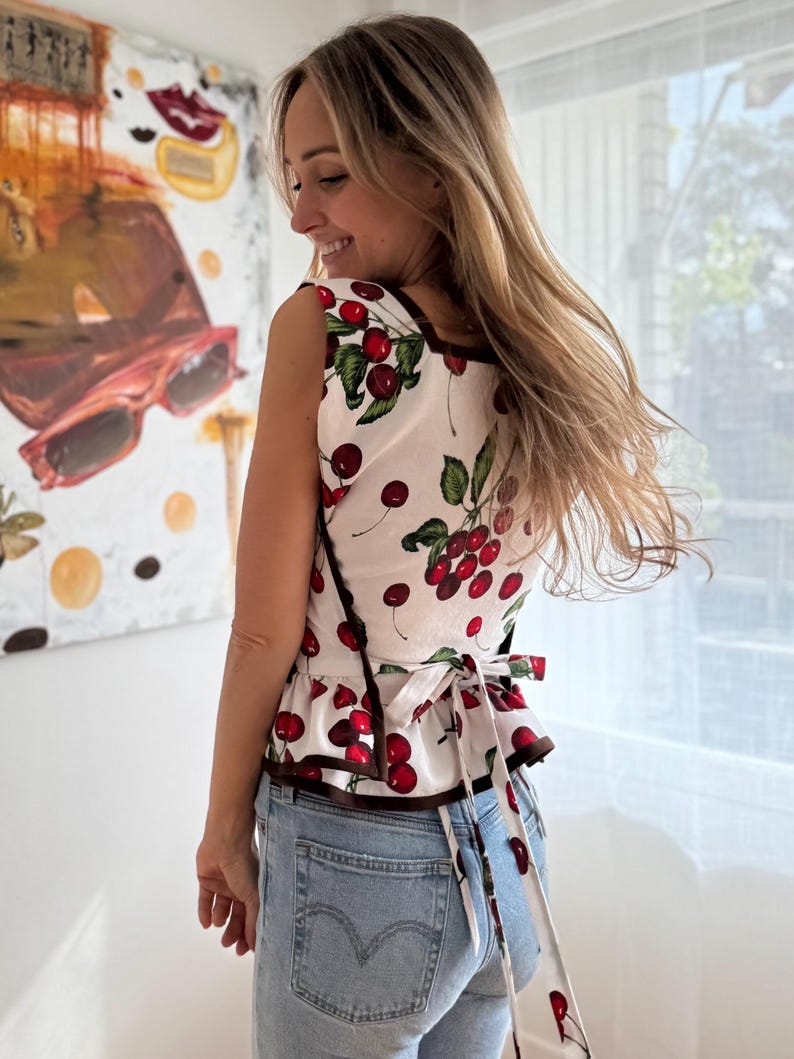 May include: A sleeveless white top with a cherry print, featuring red cherries and green leaves. The top has a brown trim and a tie-back detail. The person is wearing light blue denim jeans.