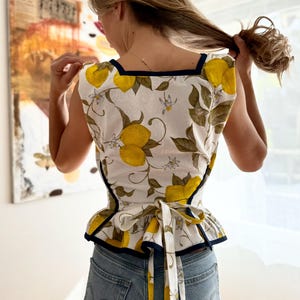 May include: A sleeveless top with a white background and a lemon pattern. The top has a square neckline with a navy blue trim and a matching tie at the waist. The lemon pattern includes yellow lemons, green leaves, and white flowers.