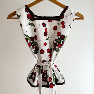 May include: A white top with a cherry print, featuring red cherries and green leaves. The top has a square neckline, brown trim, and a tie-waist design. It is hanging on a wooden hanger.