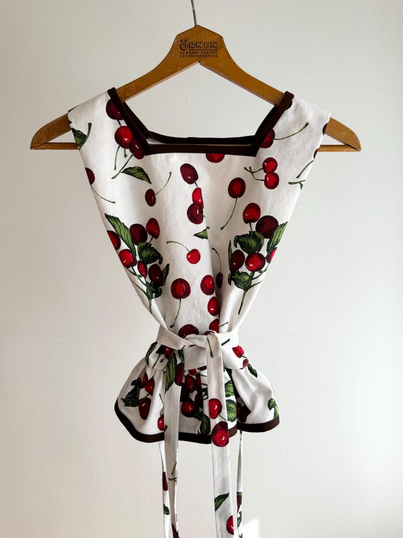 May include: A white top with a cherry print, featuring red cherries and green leaves. The top has a square neckline, brown trim, and a tie-waist design. It is hanging on a wooden hanger.