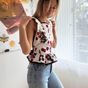 May include: A white top with a cherry print, brown trim, and a ruffled hem. The sleeveless top features a square neckline and is paired with light blue denim jeans. The top's design includes red cherries and green leaves.
