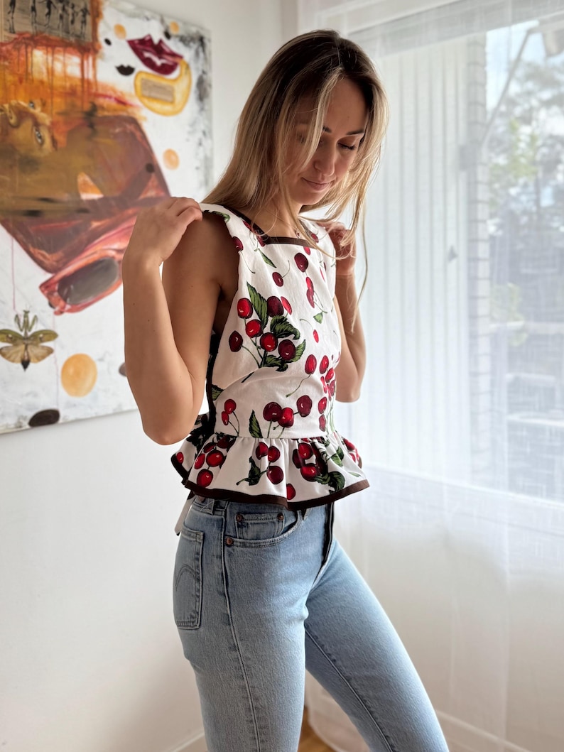May include: A white top with a cherry print, brown trim, and a ruffled hem. The sleeveless top features a square neckline and is paired with light blue denim jeans. The top's design includes red cherries and green leaves.