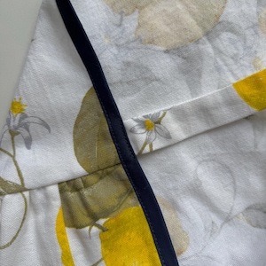 May include: White fabric with a lemon and floral pattern, trimmed with a navy blue border. The design features yellow lemons, green leaves, and small white flowers. The fabric appears to be linen or a similar natural fiber.