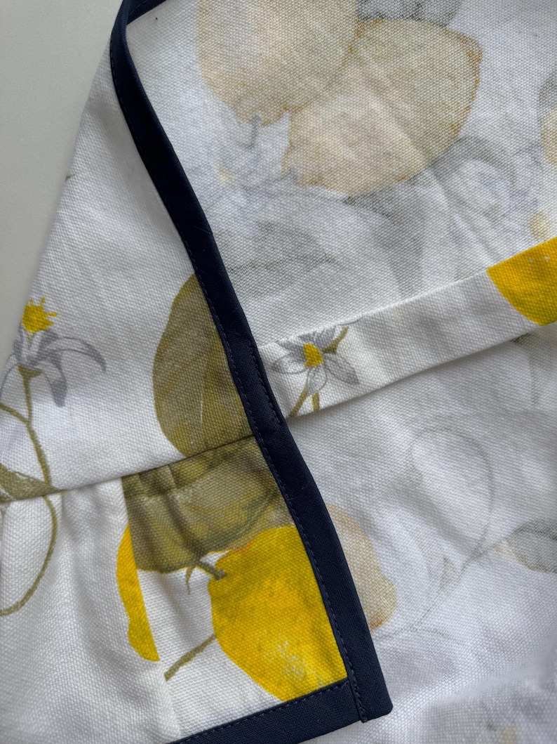 May include: White fabric with a lemon and floral pattern, trimmed with a navy blue border. The design features yellow lemons, green leaves, and small white flowers. The fabric appears to be linen or a similar natural fiber.