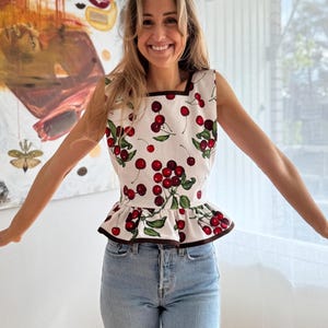 Puede incluir: Top blanco sin mangas con estampado de cerezas, con cerezas rojas y hojas verdes. El top tiene un escote cuadrado y un dobladillo con volantes con ribete marrón. La persona lleva vaqueros azul claro.