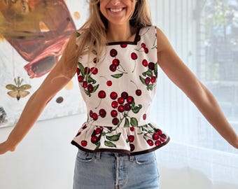 May include: A sleeveless white peplum top featuring a repeating pattern of red cherries with green leaves. The top has a square neckline and a hemline finished with a dark brown trim. The garment is paired with light blue denim jeans.